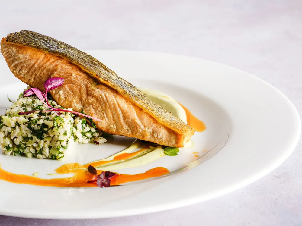 A seared salmon fillet on a white plate with a grain salad and a swirl of orange sauce
